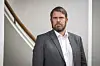 Mann i grå dress står ved en trapp med trehåndlist i et lyst innemiljø.