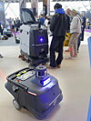 En profesjonell liten feierobot vises på stand under en renholdsmesse. I bakgrunnen ses flere mennesker som ser på en annen rengjøringsmaskin.